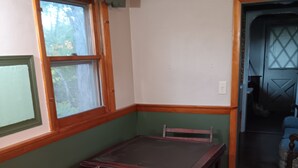 Un coin repas avec une table et des chaises en bois, une fenêtre agrémentée d’un lambrequin vert et une porte donnant sur une autre pièce.