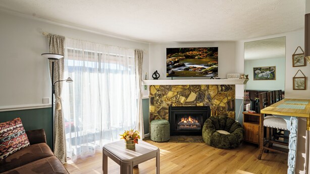A living room with a stone fireplace, a television mounted above it, a sofa, a coffee table with flowers, and a dining area with a table and chairs. - Sunset Lover - Breathtaking Balcony Views, Sleeps 4, 15 Minutes Walk to Downtown (Gatlinburg)