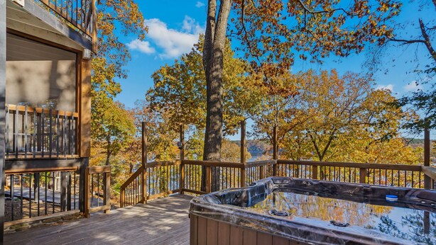 A wooden deck with a hot tub, surrounded by trees with autumn foliage, and a clear blue sky. - Dragonfly Landing on Beaver Lake, w/ hot tub (Rogers)