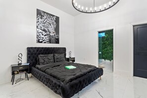 A modern bedroom with a tufted black bed, a leopard print artwork, a bedside table with a lamp, and a view of greenery through an open door.