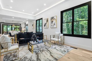 A modern living room with a black sofa, a glass coffee table, and a marble-patterned rug. There are large windows with a forest view, and artwork on the walls.