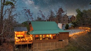 A log cabin with a green roof, a deck with lights, and a view of the night sky. - Hot tub, Sleeps 11, Near Dollywood, Pet-friendly (Sevierville)