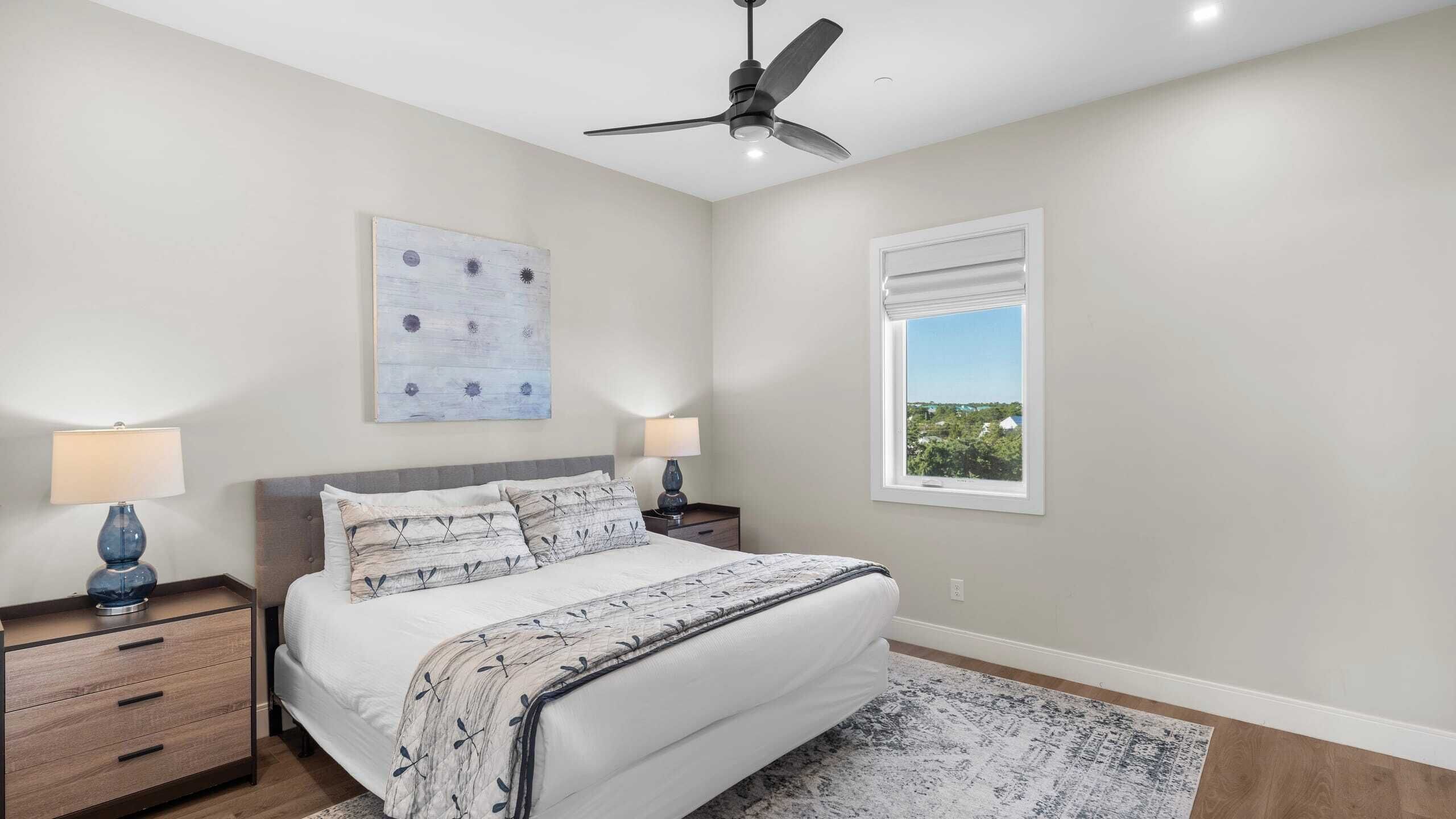 Un dormitorio con cama, dos mesitas de noche, ventilador de techo, ventana con vistas y una obra de arte colgada en la pared.