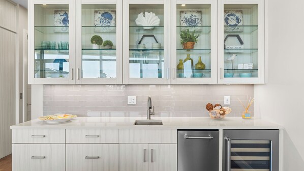 A modern kitchen with a built-in dishwasher, a stainless steel sink, and a glass-fronted cabinet displaying decorative items. - Rooftop Deck w/ Gulf Views, Game Room Modern 30A by Avantstay (Santa Rosa Beach)