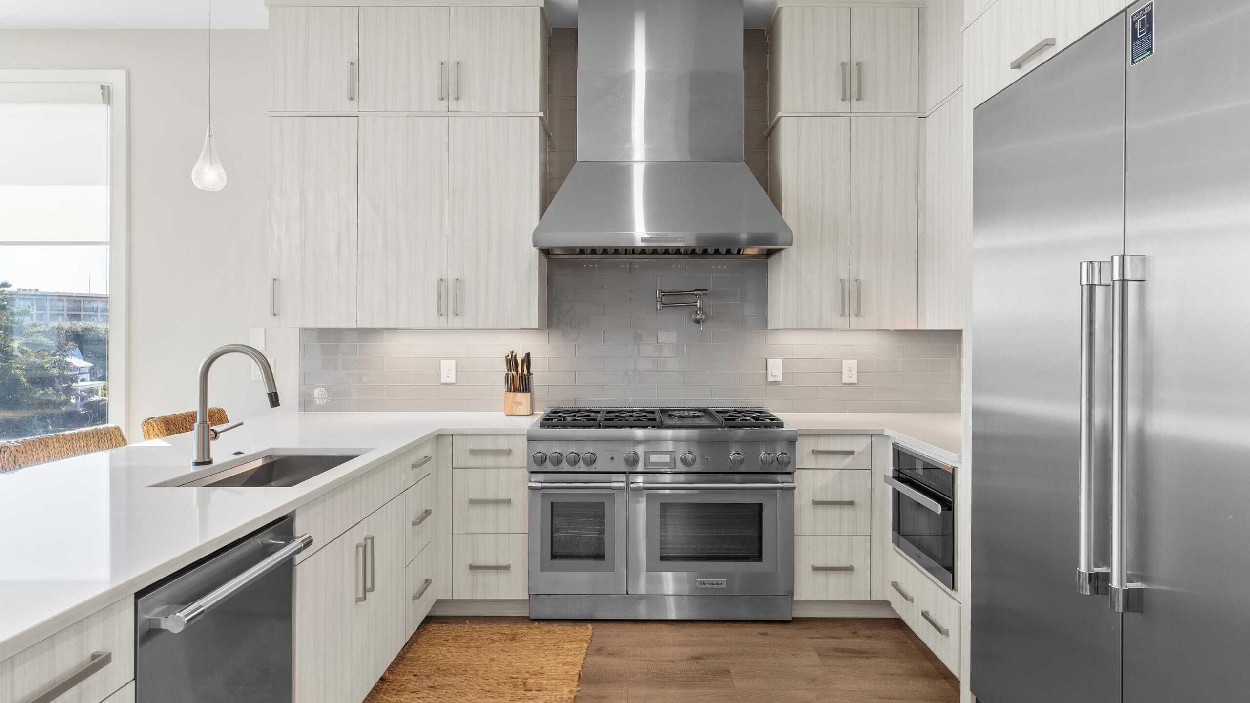 A modern kitchen with light wood cabinets, stainless steel appliances, and a large window providing a view of the city.