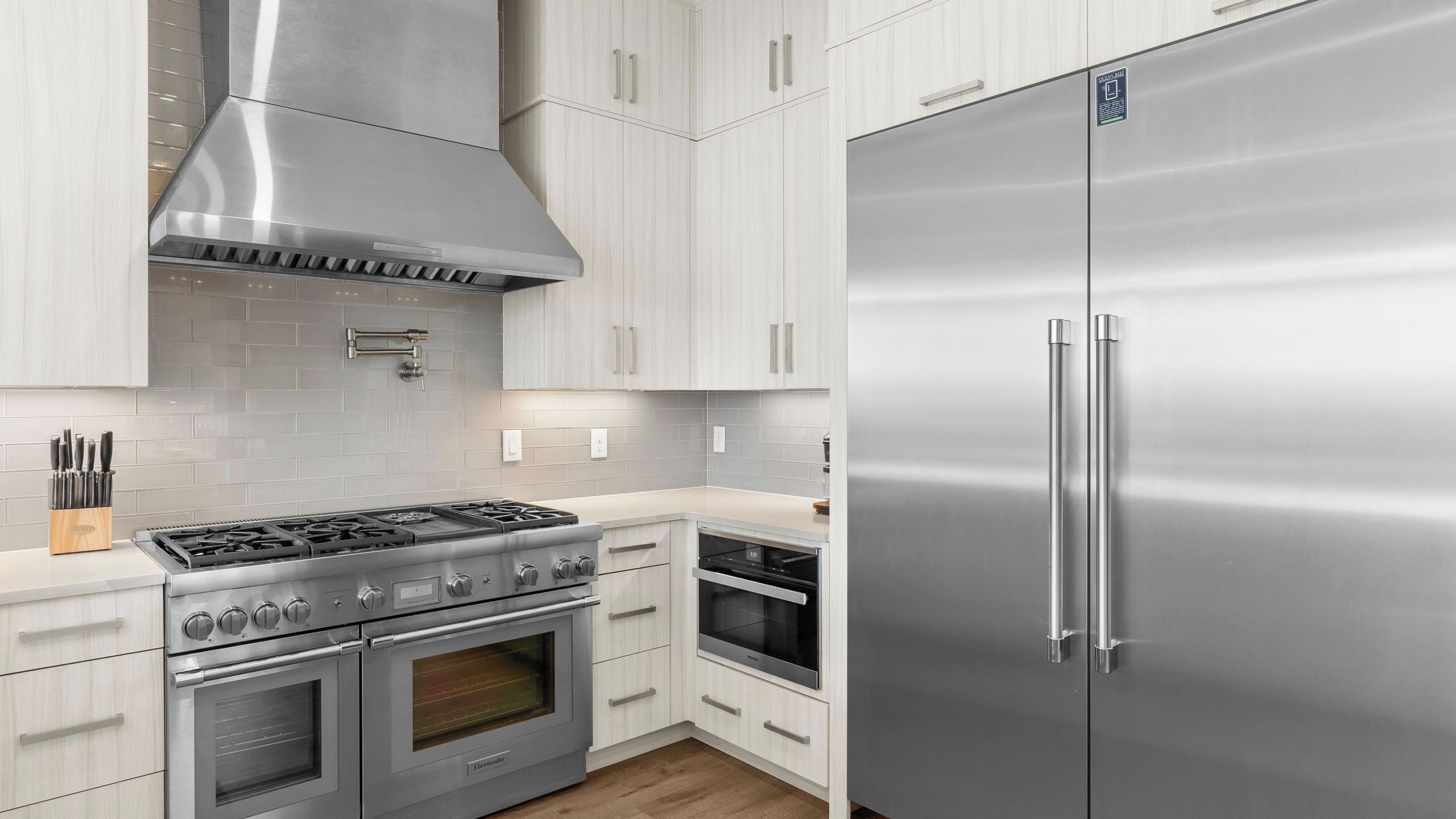 A kitchen with stainless steel appliances, including a stove, oven, and refrigerator, with light-colored cabinetry and a tiled backsplash.