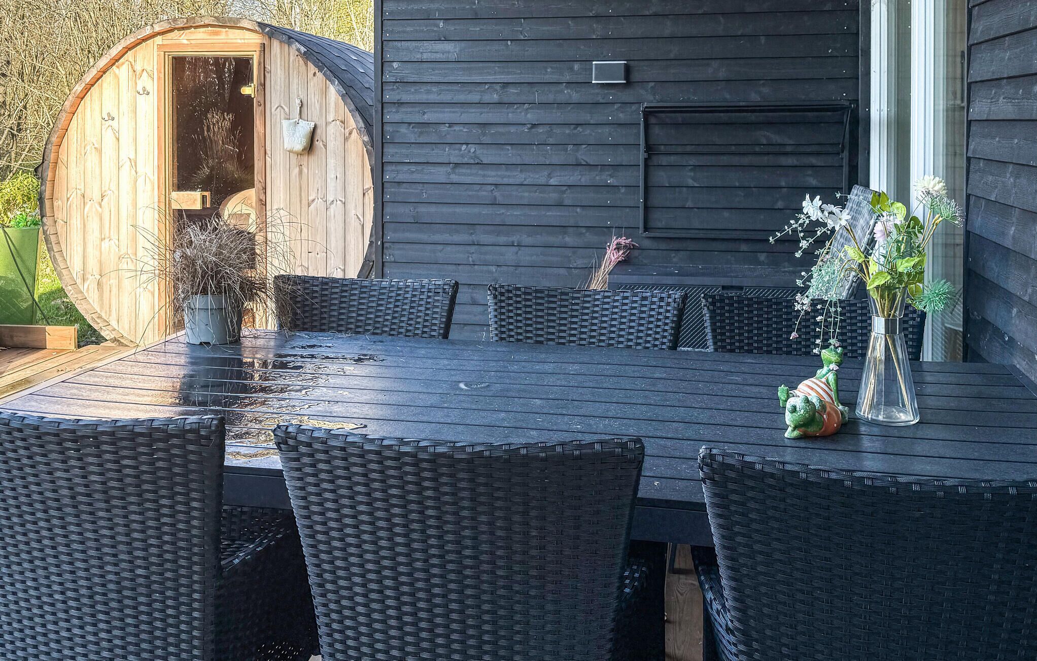 Puinen, tynnyrinmuotoinen rakennelma, jossa on ovi, tumma puinen pöytä, jonka ympärillä on punottuista materiaaleista valmistetut tuolit, sekä maljakko, jossa on kukkia.
