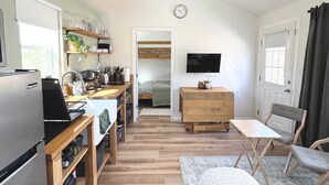 Uma cozinha compacta com geladeira, fogão e prateleiras com utensílios de cozinha. Uma área de refeições com mesa e cadeiras é visível. Uma TV está fixada na parede.