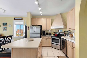 A modern kitchen with light wooden cabinets, a central island, stainless steel appliances, and a view of the outside through a window. - Maresol 310 (Ocean City)
