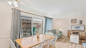 A dining area with a wooden table and white chairs, a teal sofa, and a television on a wooden cabinet. - Salty Sands 4 208 (Ocean City)