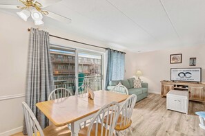 Ein Essbereich mit einem Holztisch und weißen Stühlen, einem grünen Sofa, einem Fernseher auf einer Holzkommode und einem Balkon mit Vorhängen.