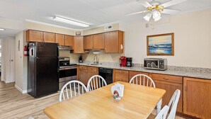 A compact kitchen with wooden cabinets, a refrigerator, microwave, and a dining table with chairs. - Salty Sands 4 208 (Ocean City)