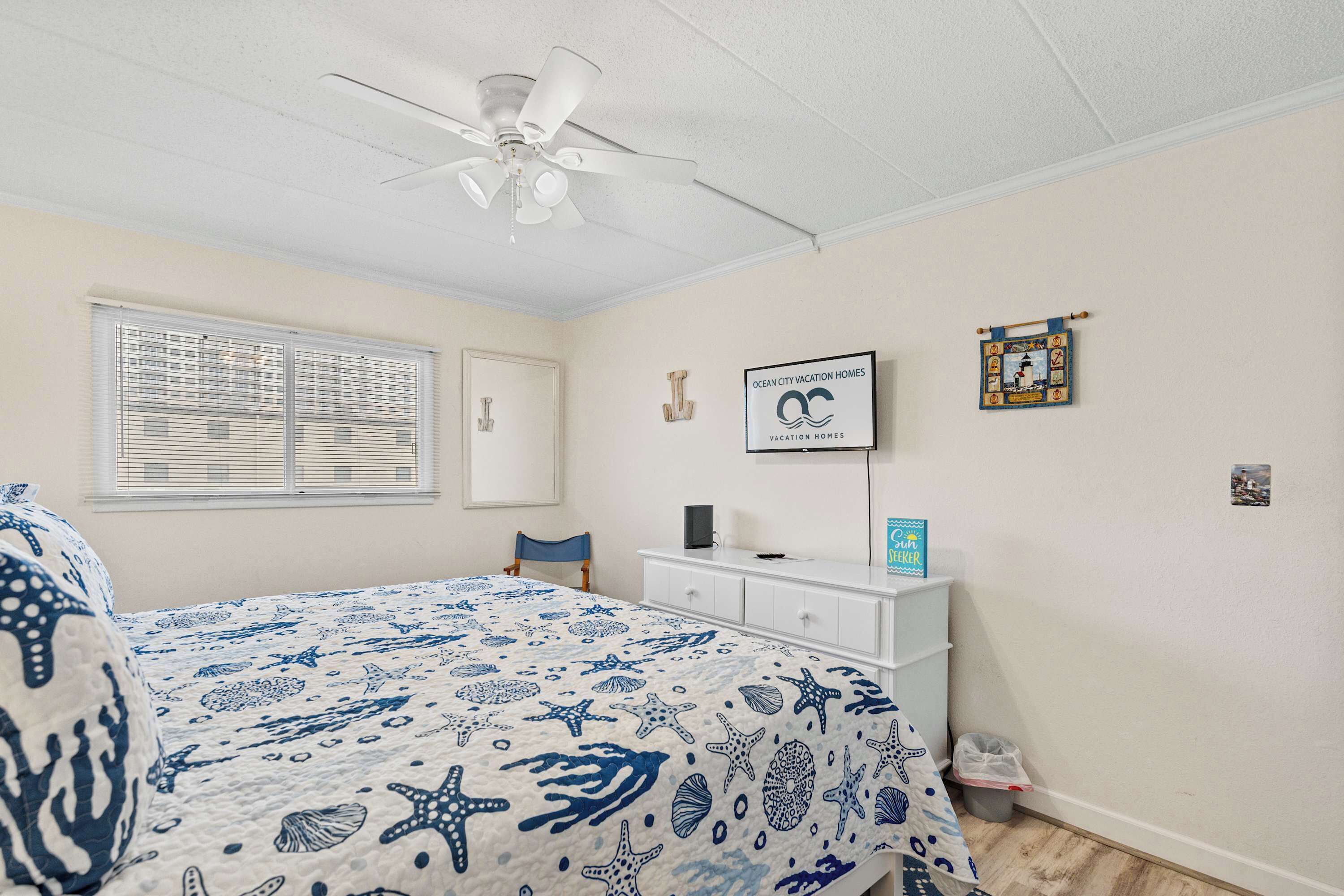 Quarto com cama, ventilador de teto, janela com persianas, escrivaninha com computador e um quadro na parede.