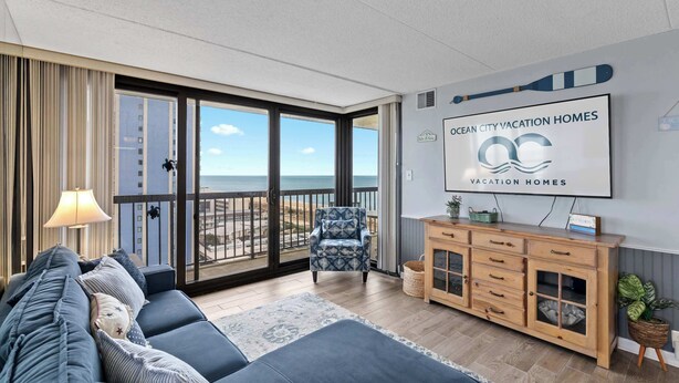 Ein geräumiges Wohnzimmer mit großem Fenster und Blick auf den Strand, einer gemütlichen Couch, einem Holzschrank und einem Fernseher, der das Logo von Ocean City Vacation Homes zeigt.