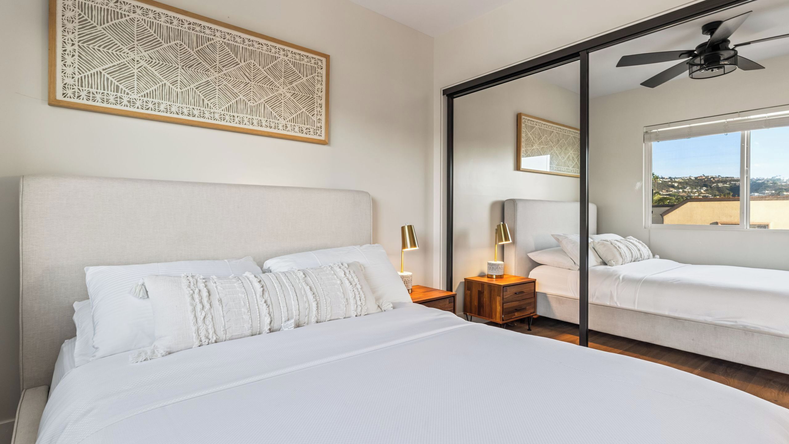 A bedroom with a large bed, a ceiling fan, a nightstand with a lamp, and a window with a view.