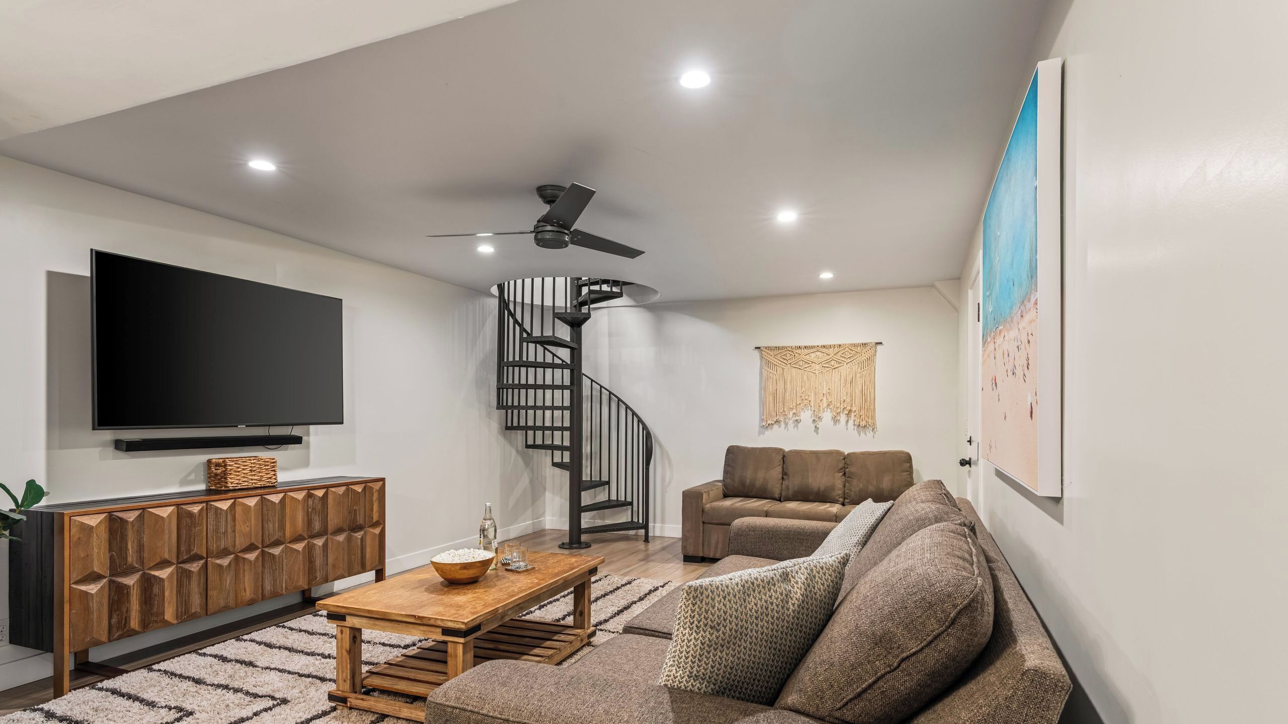 A modern living room with a flat-screen TV, a wooden coffee table, a brown sofa, a spiral staircase, and a ceiling fan.