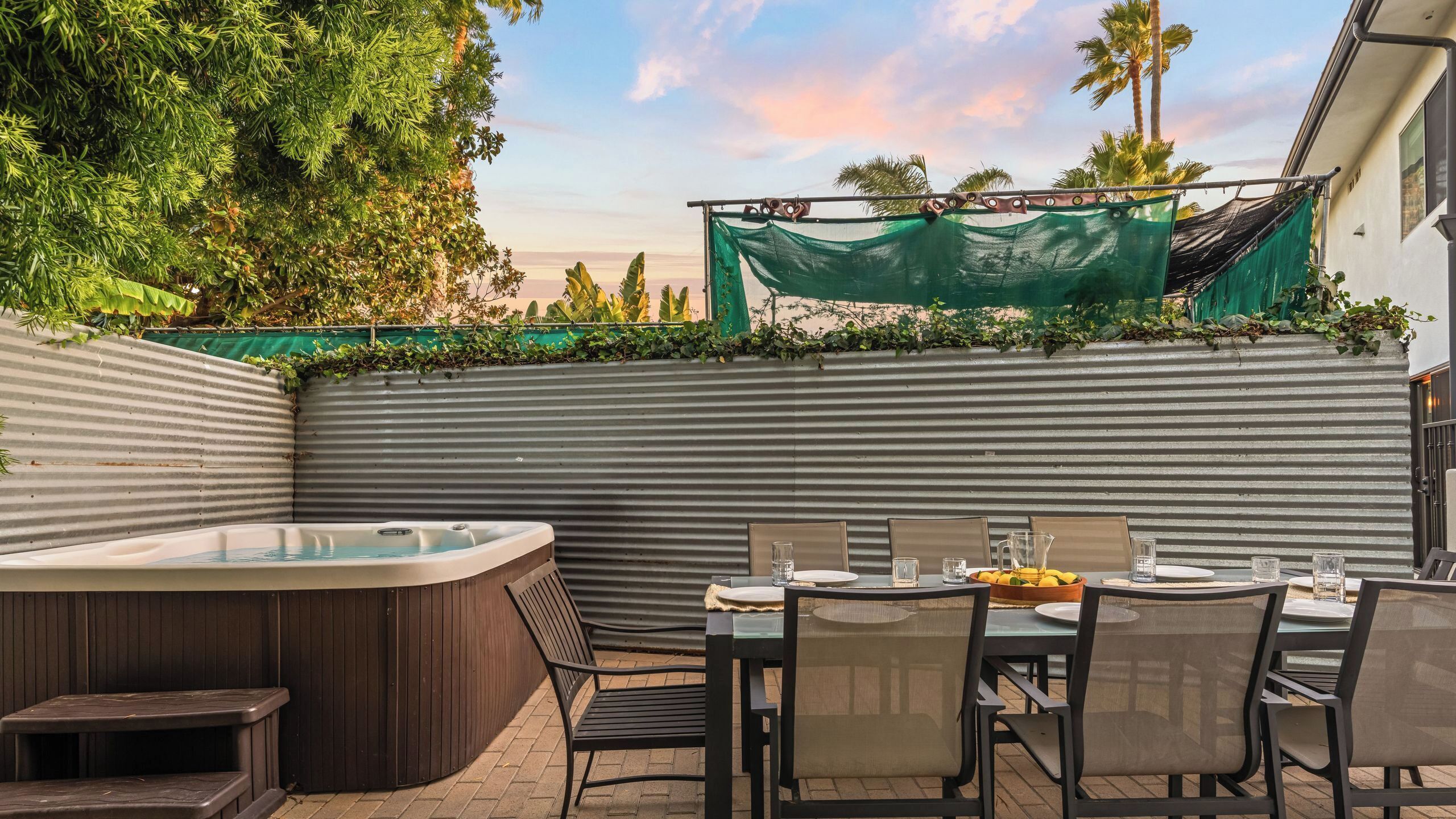 An outdoor patio with a hot tub, dining table, chairs, and a view of palm trees.