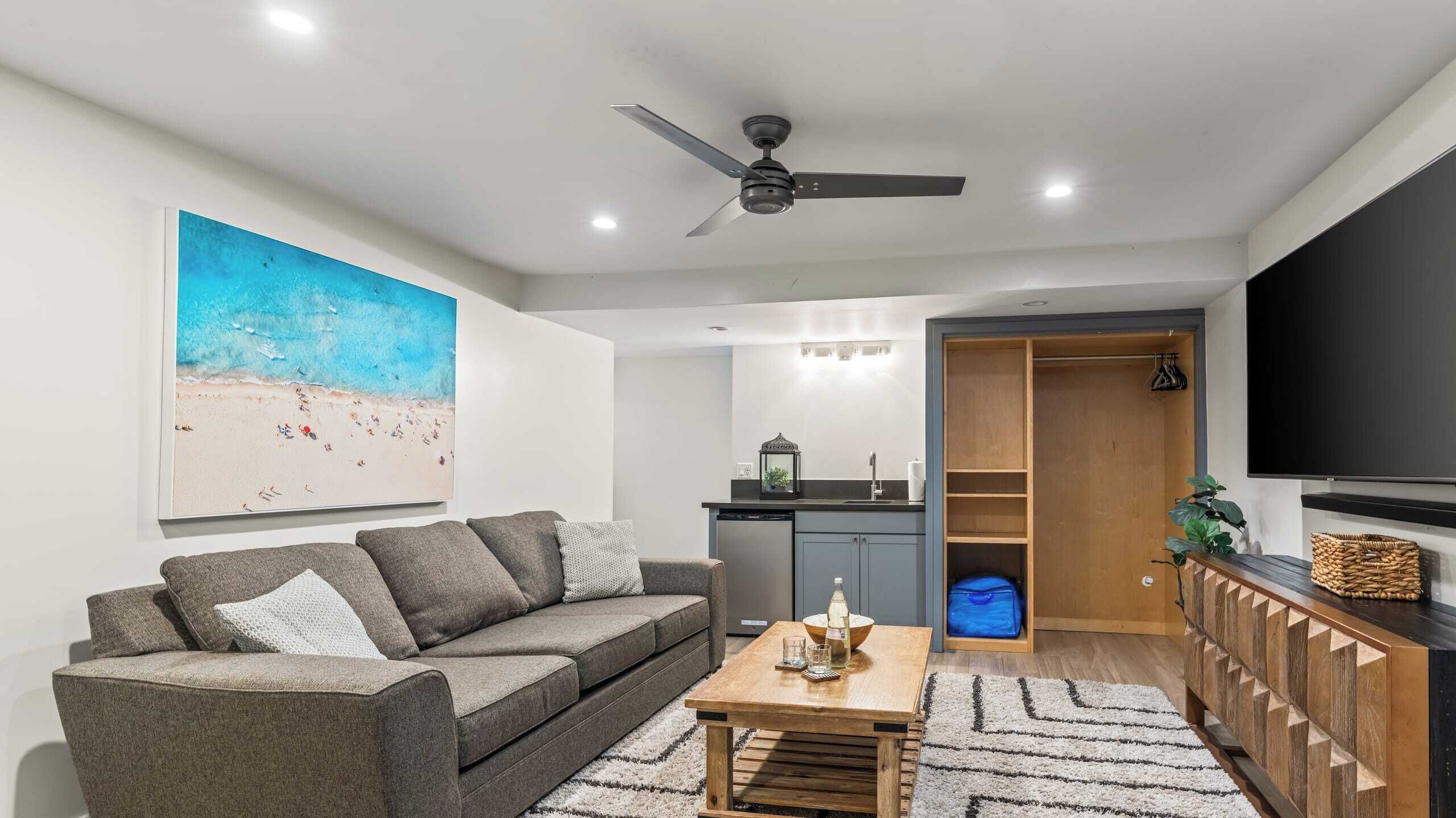 A living room with a gray sofa, a coffee table, a ceiling fan, a kitchen area, and a TV on the wall.