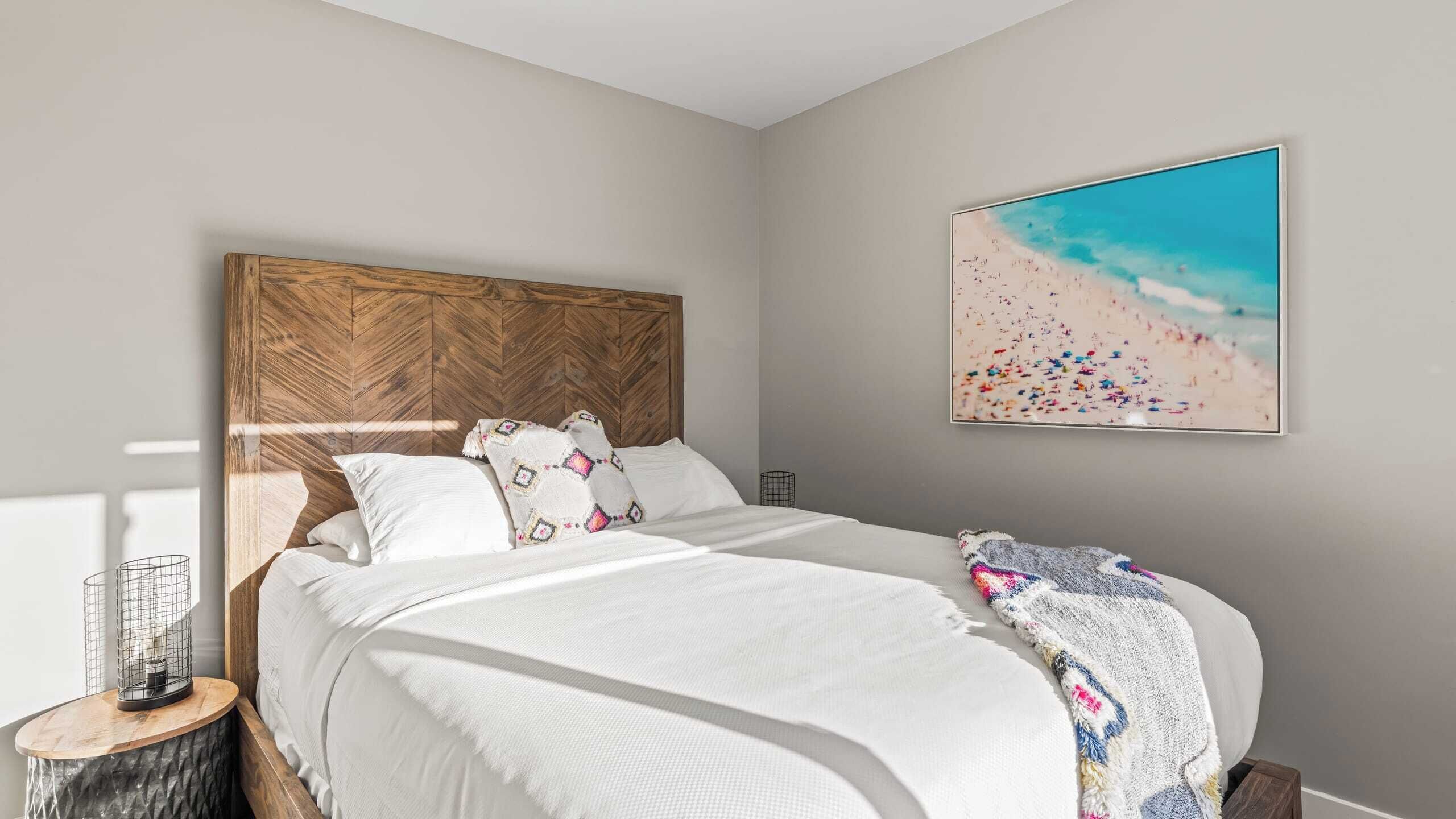 Une chambre à coucher avec une tête de lit en bois, un grand lit avec une literie blanche, un tableau sur le thème de la plage accroché au mur, et une petite table ronde sur laquelle se trouve un panier en fil de fer.