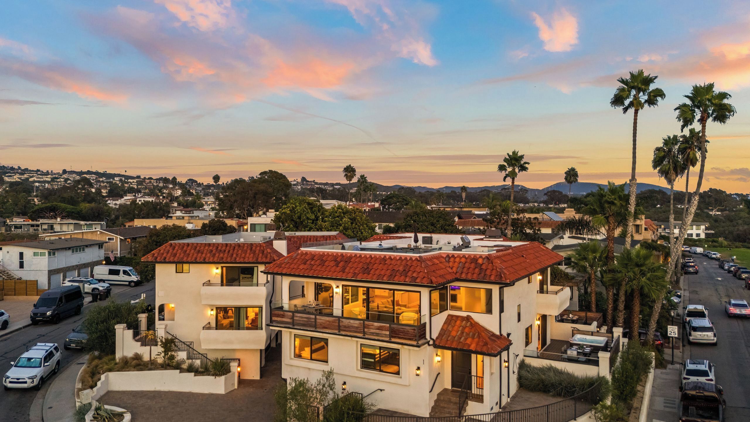 A multi-story residential building with a red-tiled roof, multiple balconies, and a view of a residential area with palm trees and distant hills.
