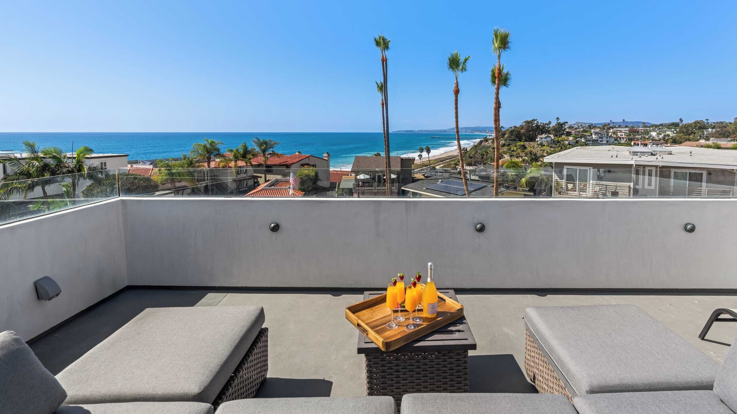 A rooftop terrace with a view of the ocean, furnished with grey sofas and a wooden table with drinks.