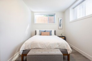Une chambre à coucher comprenant un lit, une table de chevet, une fenêtre avec des stores et un tableau accroché au mur.