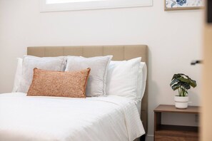 Un lit avec une literie blanche et un seul oreiller marron, une petite table de chevet avec une plante, et un tableau encadré au mur.