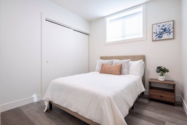 Un dormitorio con cama, mesita de noche, planta en maceta, ventana con persianas y una puerta.