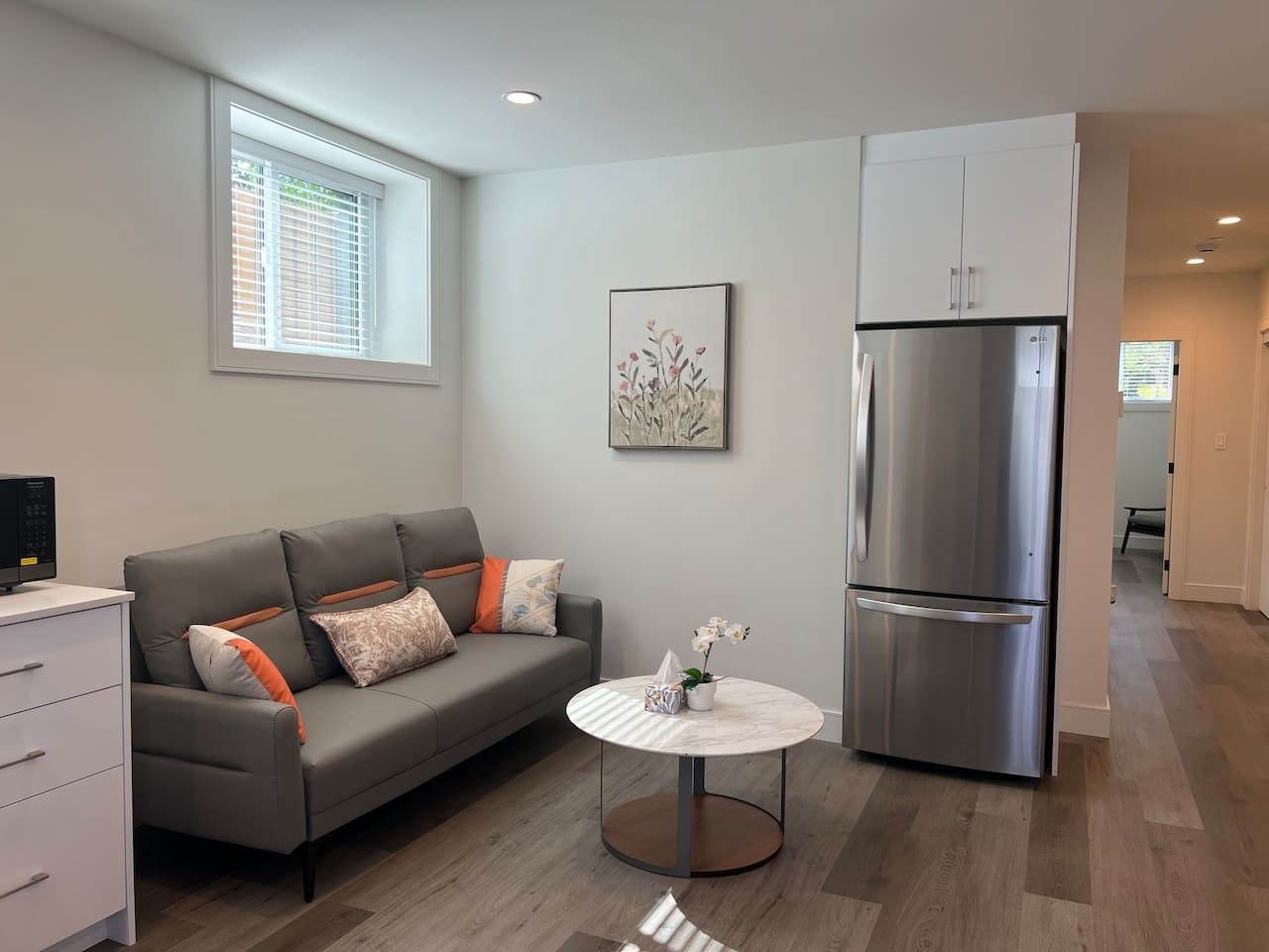 A modern living room with a grey sofa, a small round table with a vase, a stainless steel refrigerator, and a window with blinds.