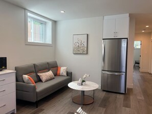 Una sala de estar moderna con un sofá gris, una mesita redonda con un florero, un refrigerador de acero inoxidable y una ventana con persianas.