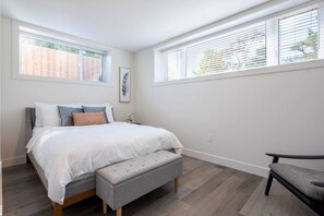 A bedroom with a bed, bench, and chair, large windows with blinds, and a wall-mounted light switch.