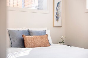 Una cama bien hecha con ropa de cama blanca y dos almohadas, una azul y otra estampada, en una habitación con una ventana y una obra de arte enmarcada en la pared.