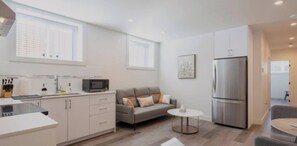 Una cocina moderna con microondas, pileta y alacenas, un sofá gris con almohadones, una mesita redonda y un refrigerador grande.