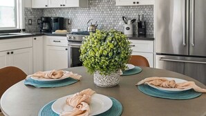 A modern kitchen with white cabinets, a stainless steel refrigerator, and a round dining table with placemats and plates. - Walk to Beach Hot Tub + Oceanview Deck 259 Trailhead Trestles 2 by Avantstay (San Clemente)