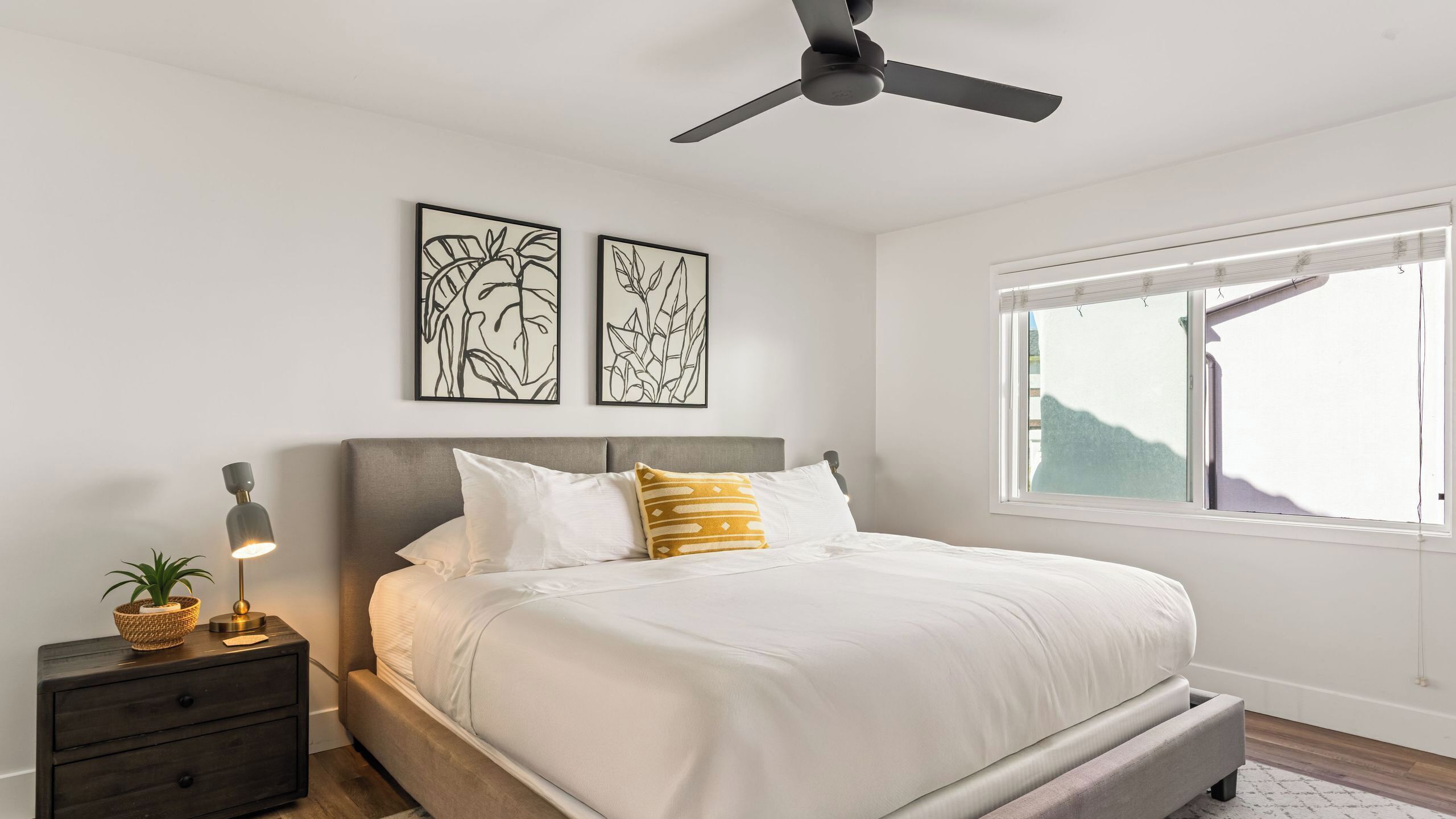 A bedroom with a bed, a nightstand, a lamp, two wall art pieces, a ceiling fan, and a window.