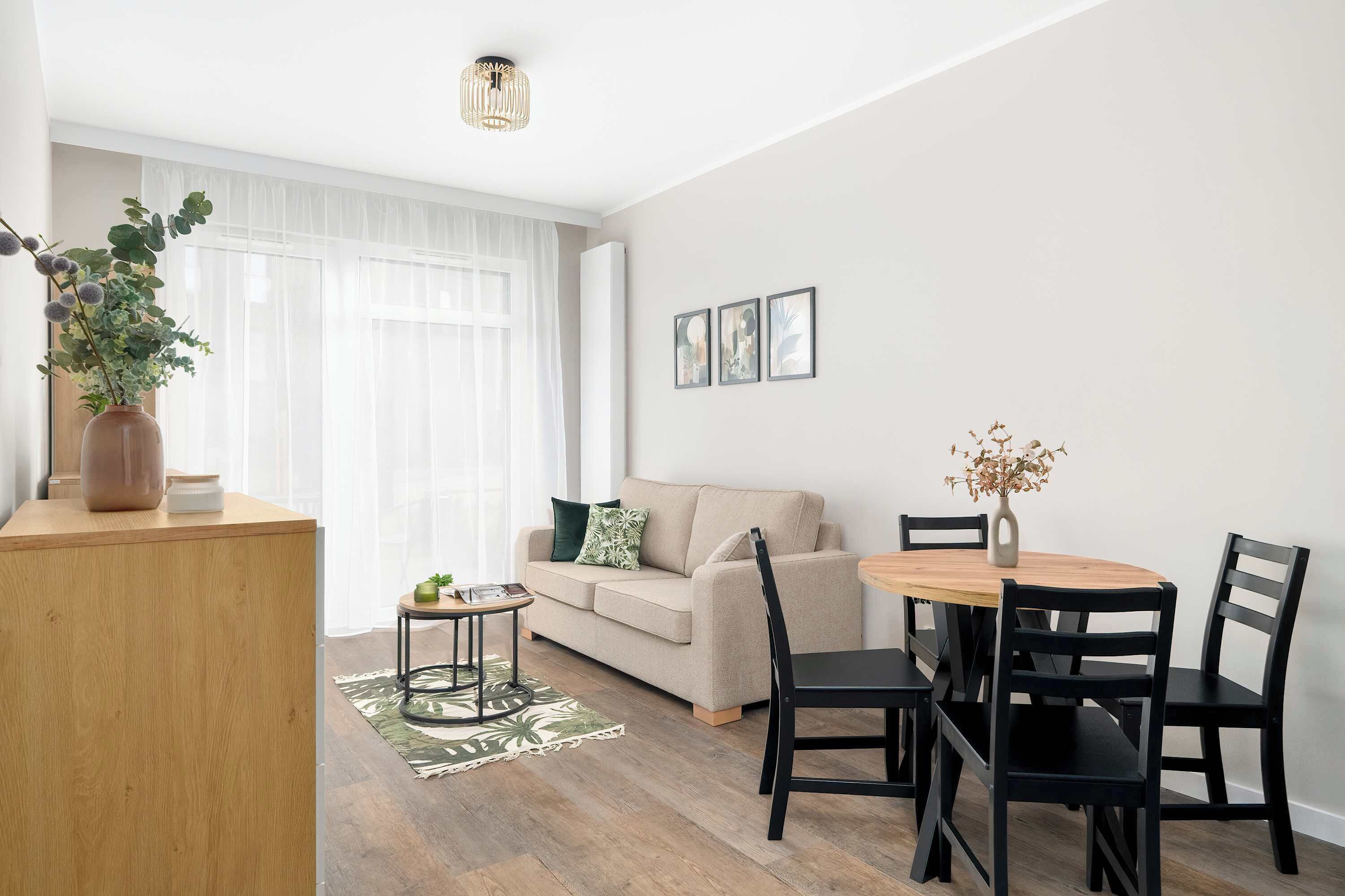 Ein modernes Wohnzimmer mit einem beigefarbenen Sofa, einem runden Holzesstisch mit schwarzen Stühlen, einem kleinen Beistelltisch mit einer Pflanze und einem großen Fenster mit leichten Vorhängen.