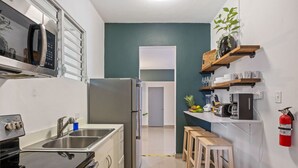 A compact kitchen with a stainless steel refrigerator, a microwave, and a sink. There are wooden stools and a shelf with kitchen items. - Ocean Park 3BD+2BA Pool Walk to beach @Portocafe (San Juan)