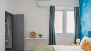 Quarto com cama, mesa de cabeceira, janela com cortinas, ar-condicionado fixado na parede e prateleira com chapéu e planta.