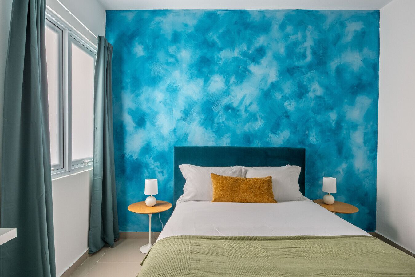 Camera da letto con un letto, due comodini, una finestra con tende e un parete azzurra con texture.