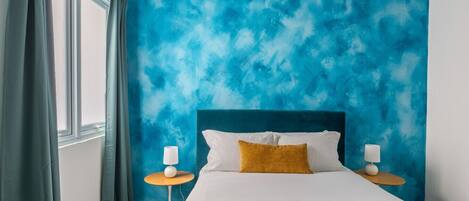 Camera da letto con un letto, due comodini, una finestra con tende e un parete azzurra con texture.