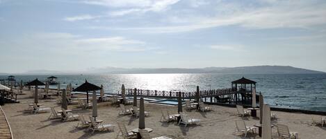 A beach with sun loungers and umbrellas, a pool, and a pier extending into the water.