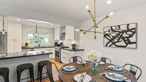 A modern kitchen with white cabinets, a central island, and a dining area with a round table set for six. - Steps to Beach, Hot Tub + Private Patio 259 Trailhead Trestles 3 by Avantstay (San Clemente)
