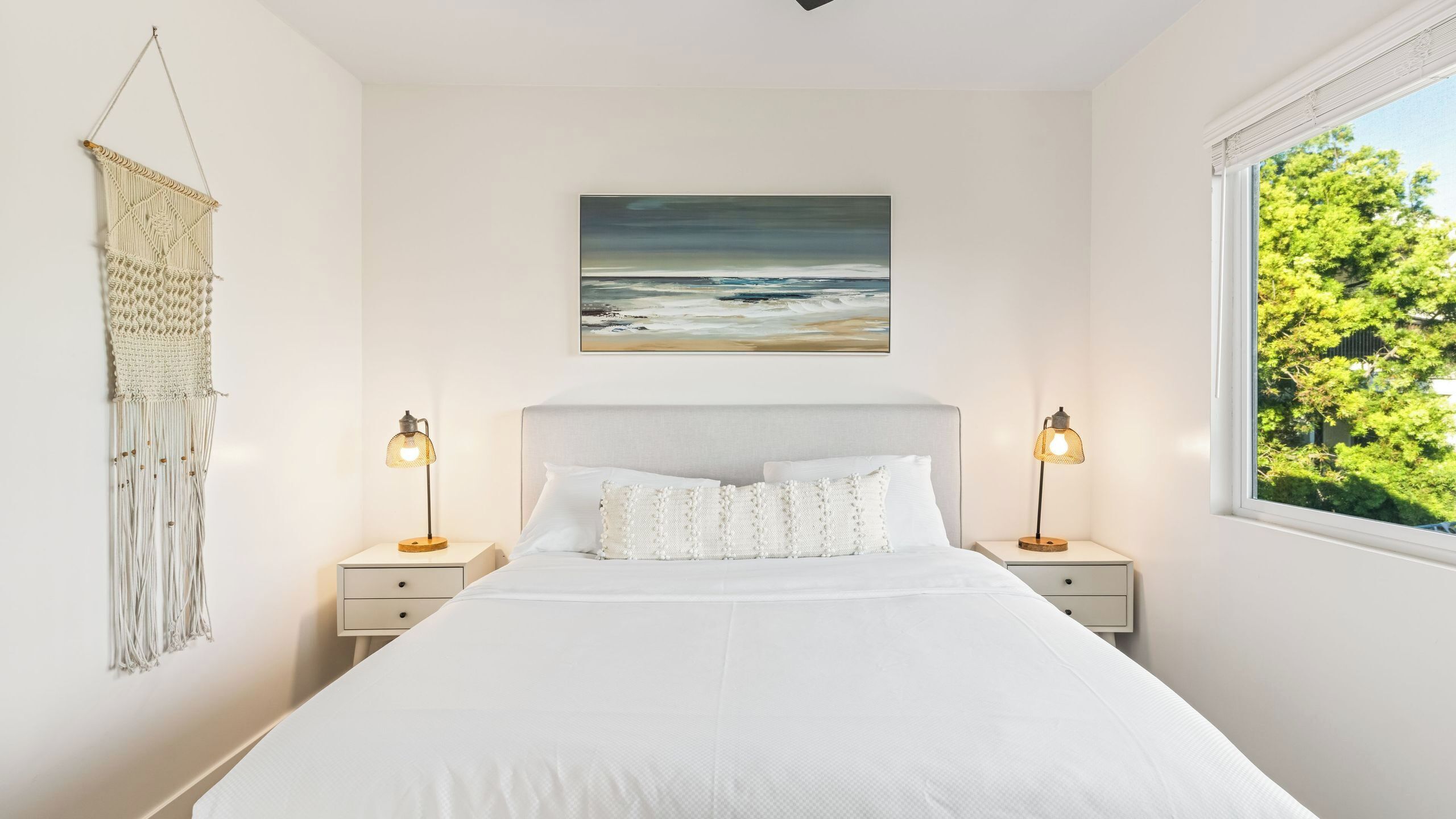 A bedroom with a bed, two bedside tables, a wall-mounted macrame, a painting of the ocean, and a window with a view of trees.
