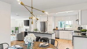 A modern kitchen with white cabinetry, a stainless steel refrigerator, and a dining area with a wooden table and chairs. - Steps to Beach, Hot Tub + Private Patio 259 Trailhead Trestles 3 by Avantstay (San Clemente)