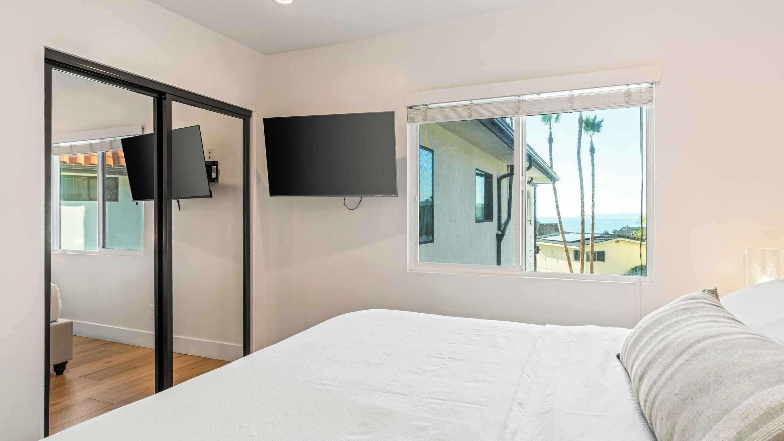 Une chambre à coucher avec un lit, un téléviseur fixé au mur et une fenêtre donnant sur des palmiers et un immeuble.