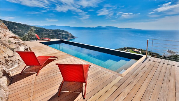 Eine Holzterrasse mit Pool und roten Stühlen mit Blick auf eine Küstenlandschaft.