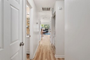 Un pasillo estrecho con paredes blancas y piso de madera, que conduce a un comedor con una mesa y sillas.