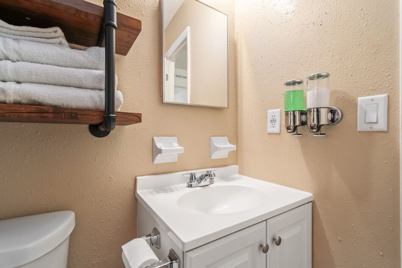 Ein Badezimmer mit einem weißen Waschbecken, einer Toilette und einem Spiegel. Handtücher liegen auf einer Holzablage und eine Seifenspender ist an der Wand angebracht.