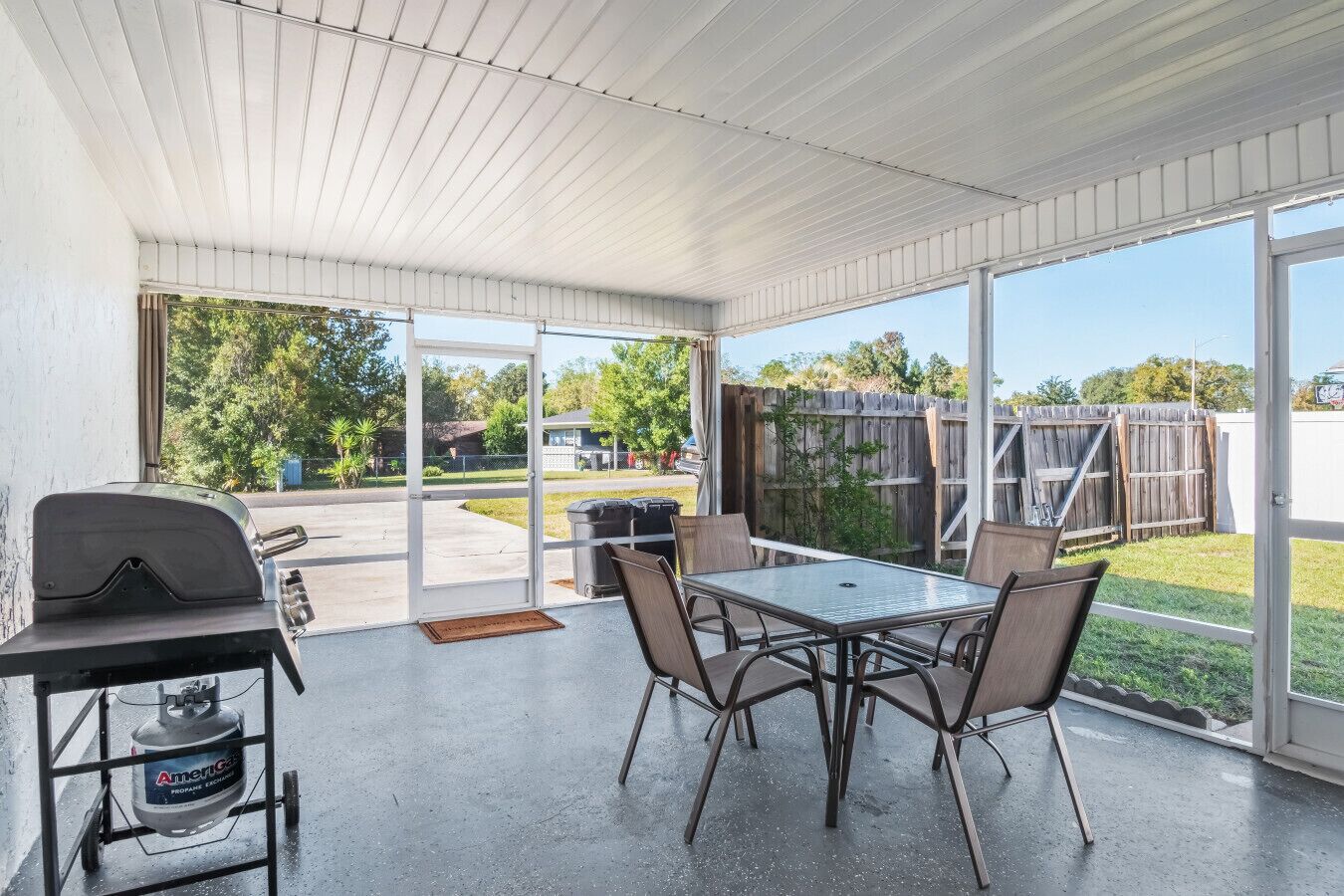 Overdekt terras met een barbecue, tafel en stoelen, met uitzicht op een achtertuin met houten omheining en begroeiing.