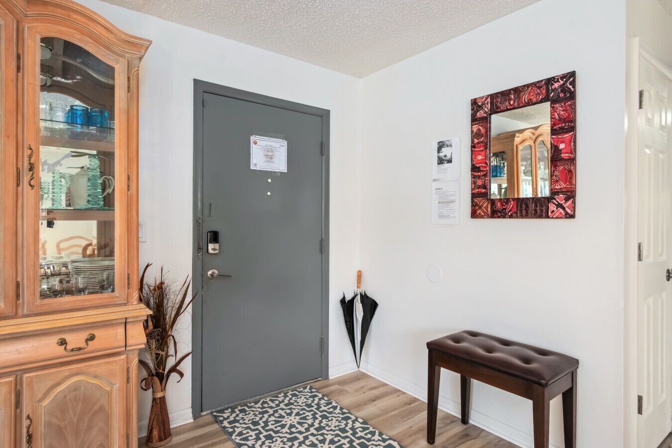 A hallway with a grey door, a wooden cabinet with glass doors, a bench, a patterned rug, and a mirror with a red frame.